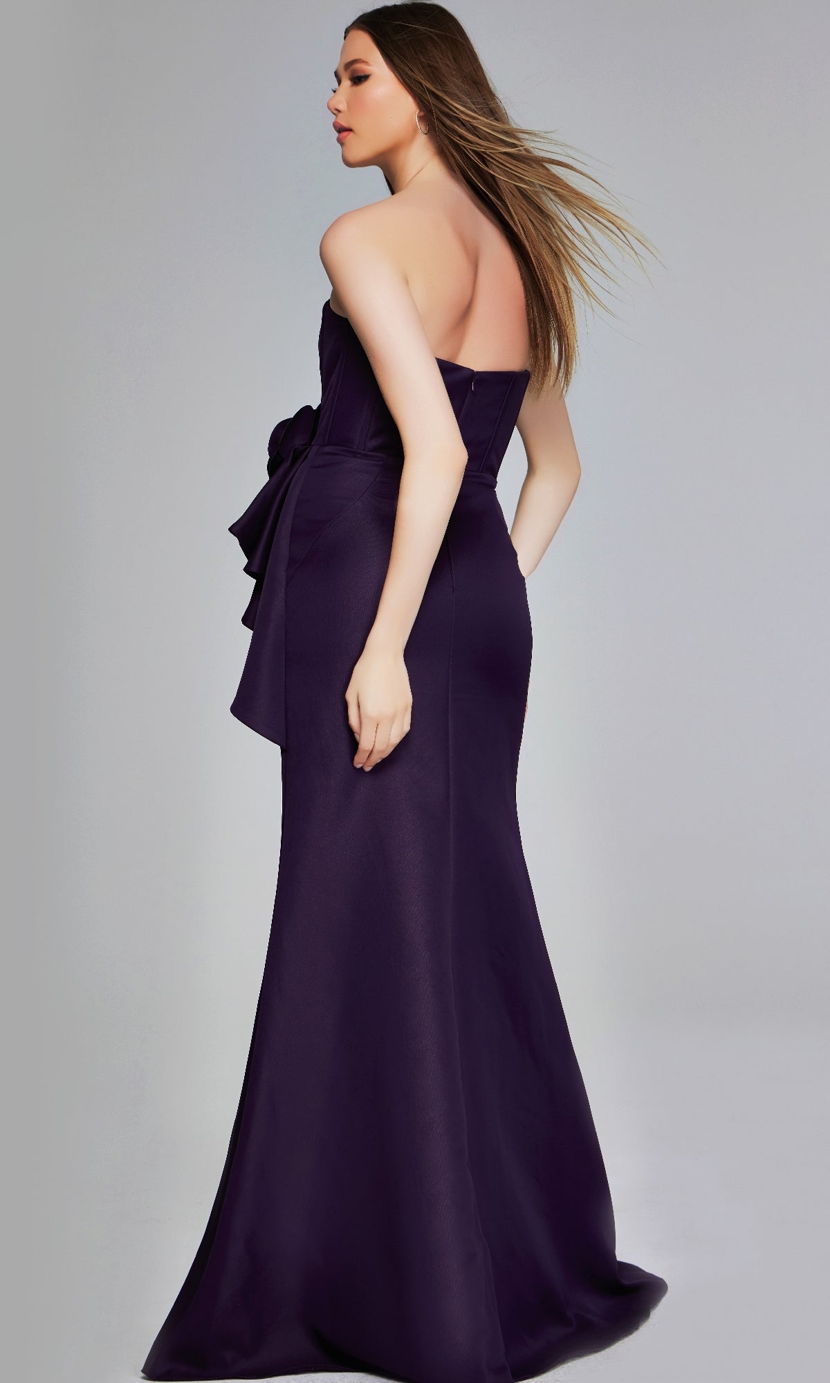 A woman faces away, wearing the Jovani 40592 long formal dress—a dark purple strapless gown with a fitted bodice, side bow detail, and floor-length silhouette.